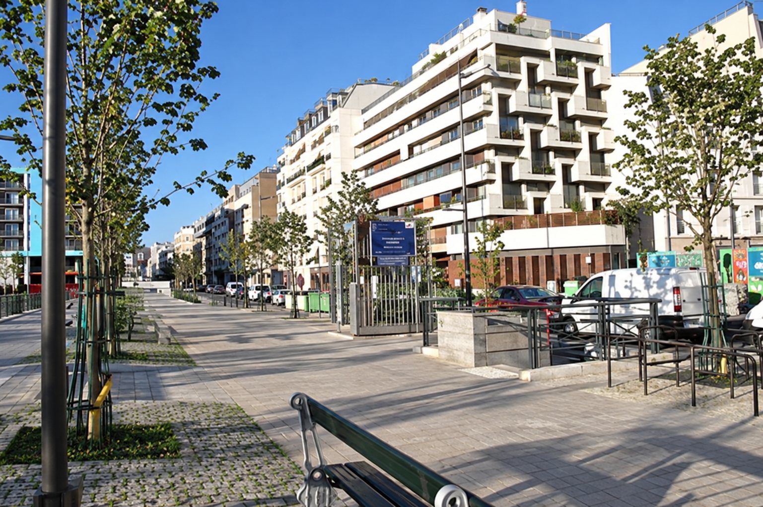 Une large avenue piétonne bordée de jeunes arbres, de bancs et d'immeubles d'habitation modernes sous un ciel bleu.