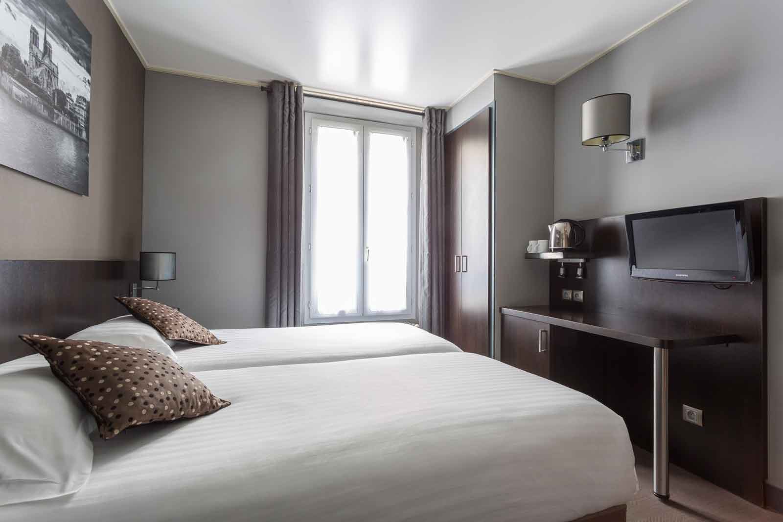Chambre d'hôtel lumineuse aux murs gris avec deux lits blancs, coussins à pois marron, bureau en bois foncé, télévision et bouilloire, et un tableau de Notre-Dame au mur.