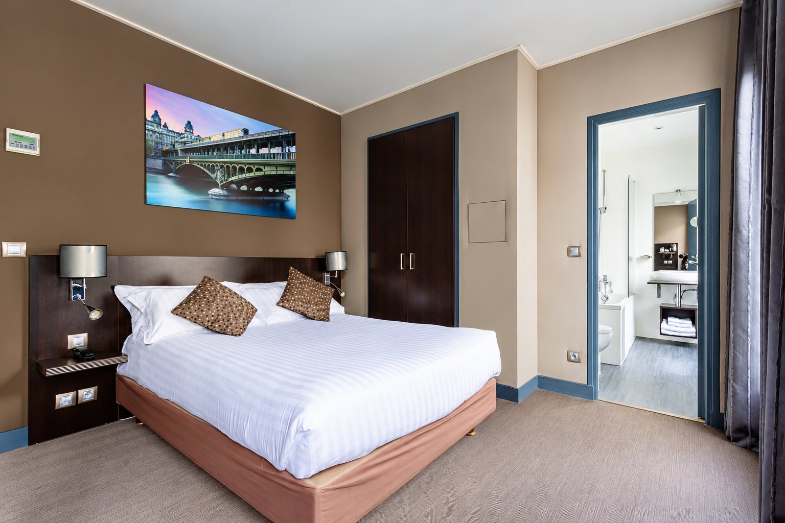 Chambre d'hôtel aux murs taupe avec un grand lit blanc, un tableau de pont parisien au-dessus et une salle de bain visible par la porte ouverte.