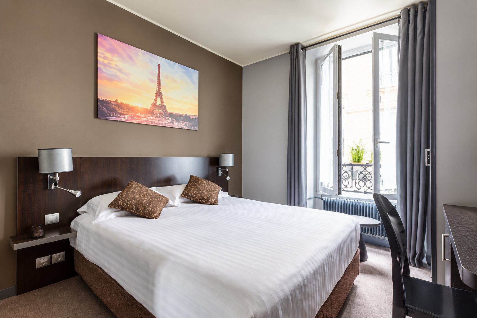 Chambre d'hôtel aux murs gris avec un grand lit blanc, un tableau de la tour Eiffel et une fenêtre ouverte avec rideaux gris.