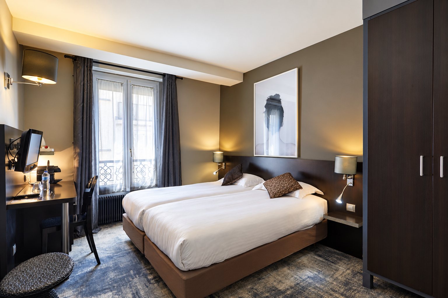 Chambre d'hôtel aux tons bruns avec deux lits jumeaux, un bureau avec télévision, une fenêtre aux rideaux gris et une armoire en bois sombre.