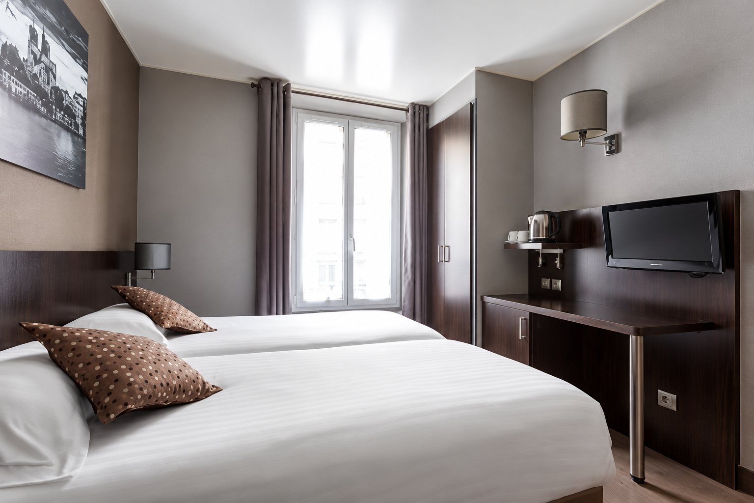 Chambre d'hôtel moderne aux tons gris et bruns avec deux lits jumeaux dressés de blanc, une fenêtre aux rideaux gris, un bureau et une télévision murale.