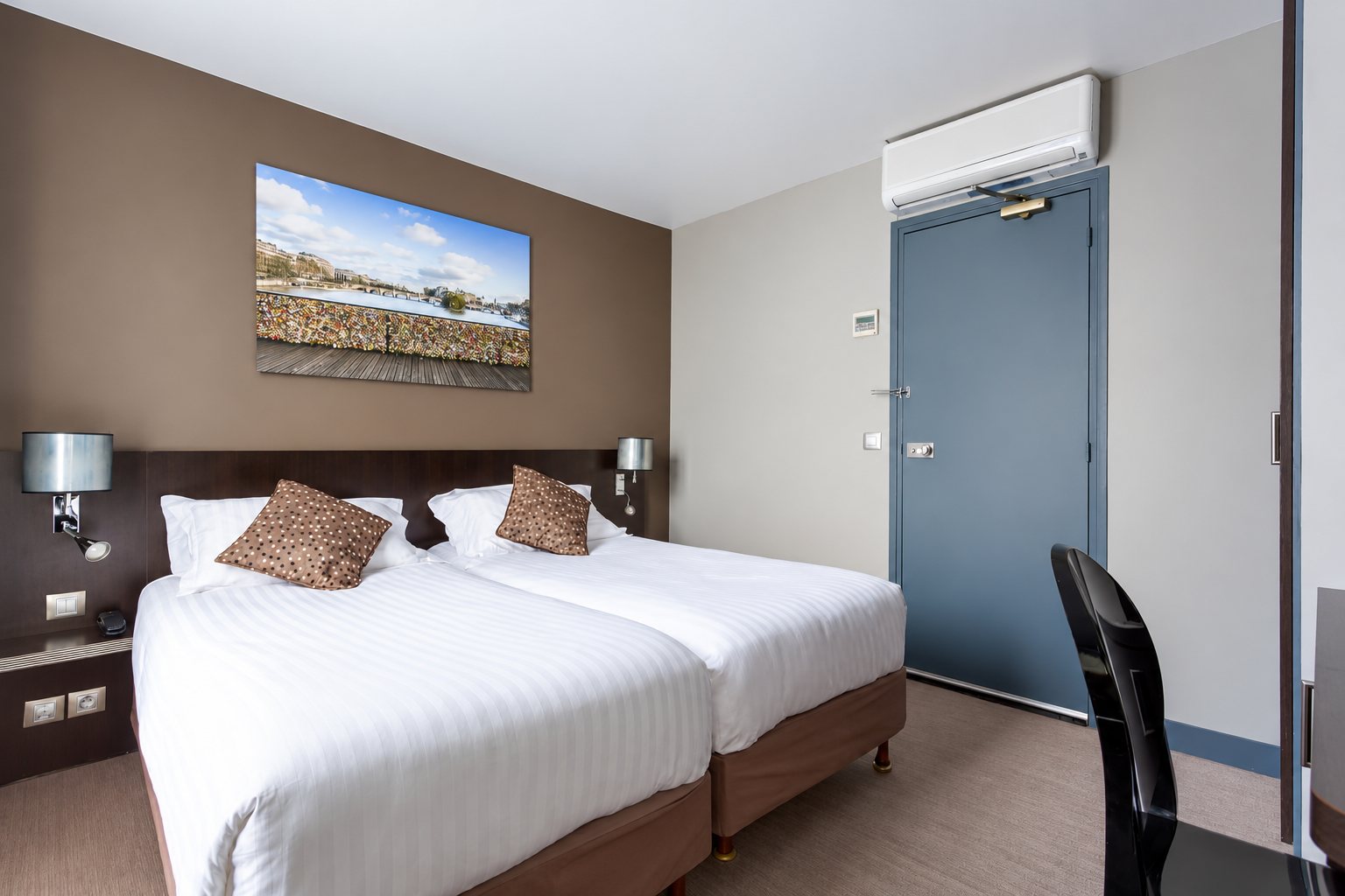 Chambre d'hôtel moderne aux murs taupe avec deux lits jumeaux dressés de linge blanc, des coussins à pois, une porte bleu-gris et un tableau d'un pont parisien au-dessus de la tête de lit.