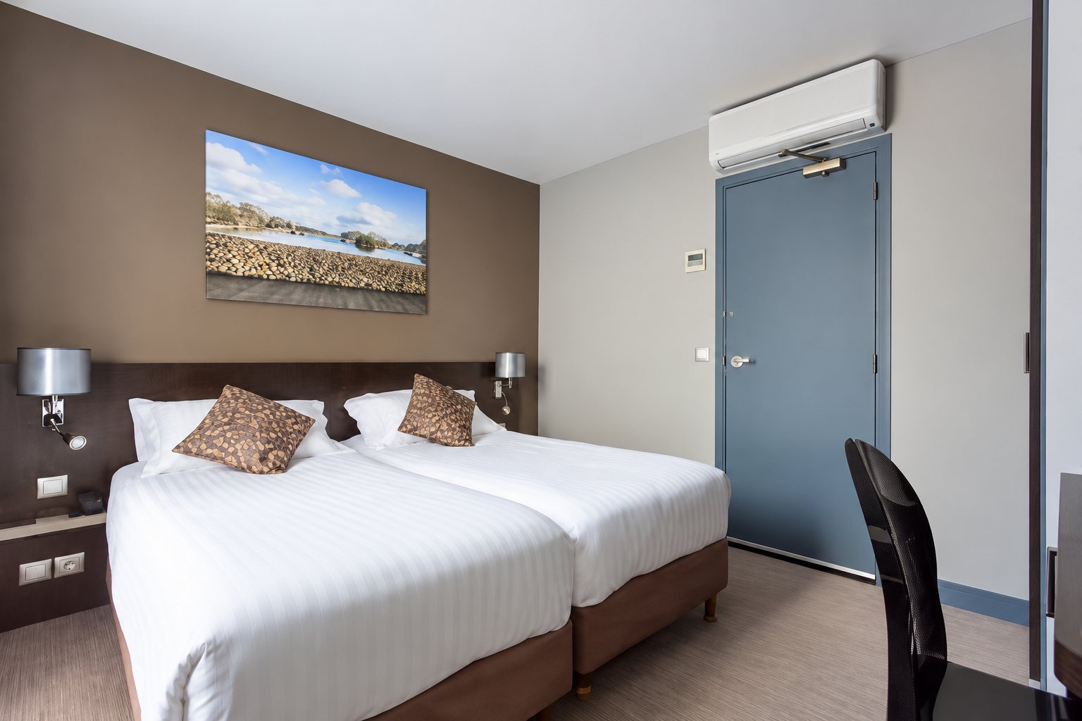 Chambre d'hôtel lumineuse avec deux lits jumeaux dotés de linge blanc, un paysage encadré au mur, une climatisation et une porte grise.