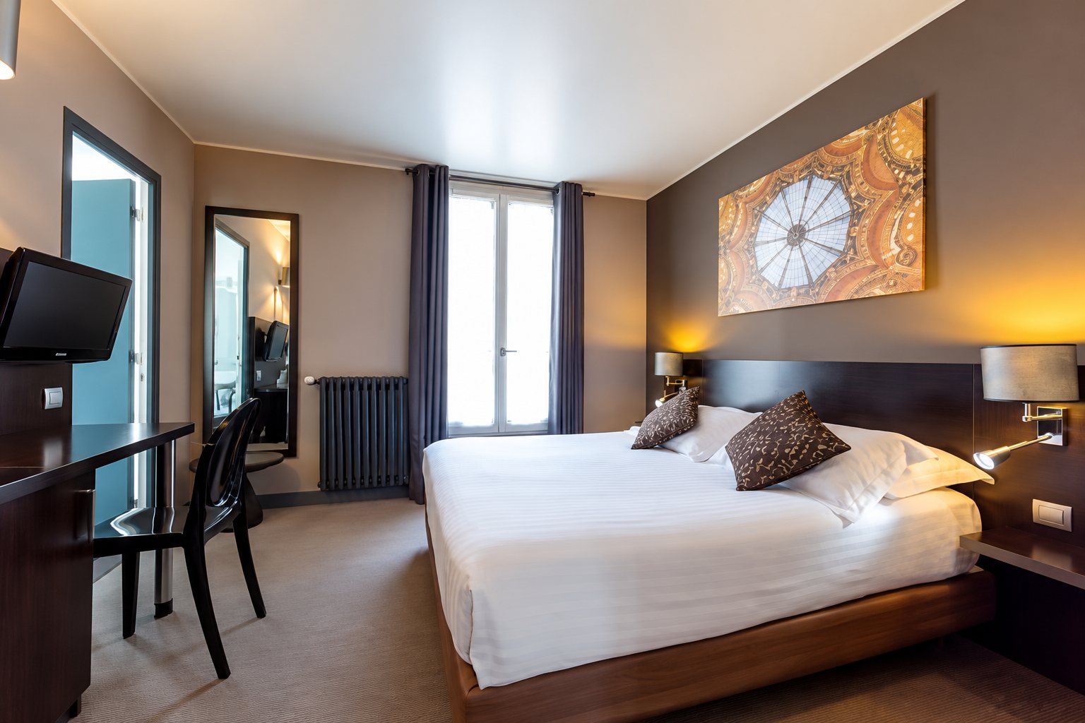 Chambre d'hôtel lumineuse avec un grand lit blanc, des coussins marron, un bureau, un téléviseur et un tableau doré au mur.