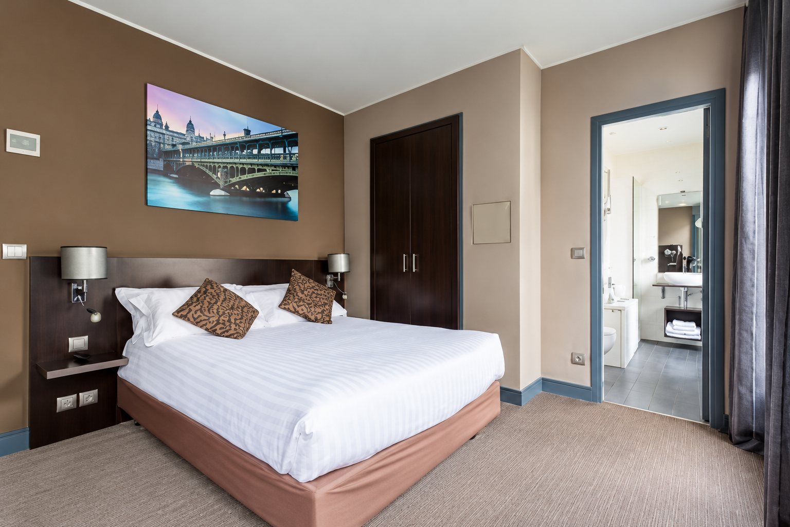 Chambre d'hôtel aux murs taupe avec un grand lit blanc, un tableau d'un pont au-dessus du lit et une salle de bain visible à droite.