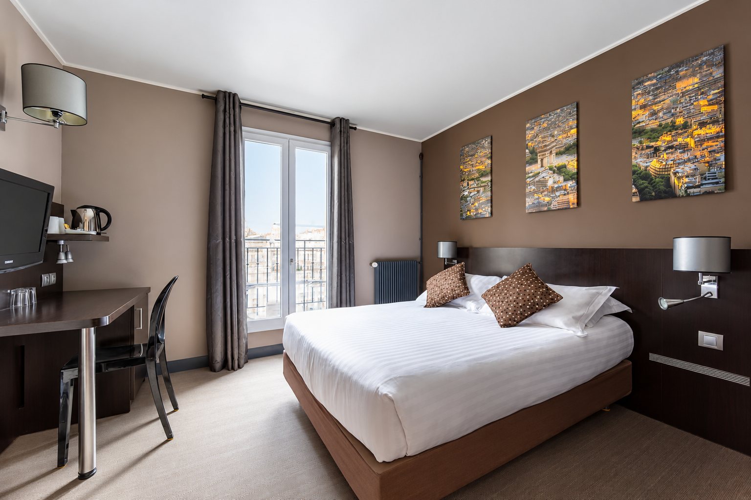 Chambre d'hôtel aux murs taupe avec un grand lit double, trois tableaux de paysages urbains, un bureau et une porte-fenêtre donnant sur un balcon.