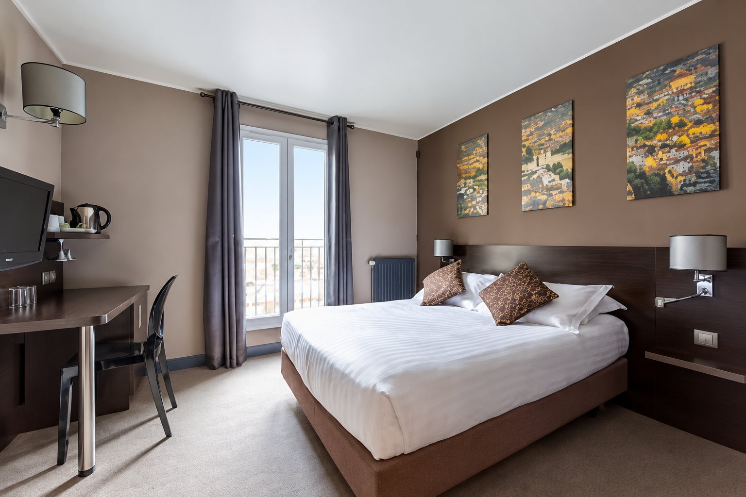 Chambre d'hôtel aux murs taupe avec un grand lit blanc, trois tableaux colorés, un bureau et une porte-fenêtre aux rideaux gris.