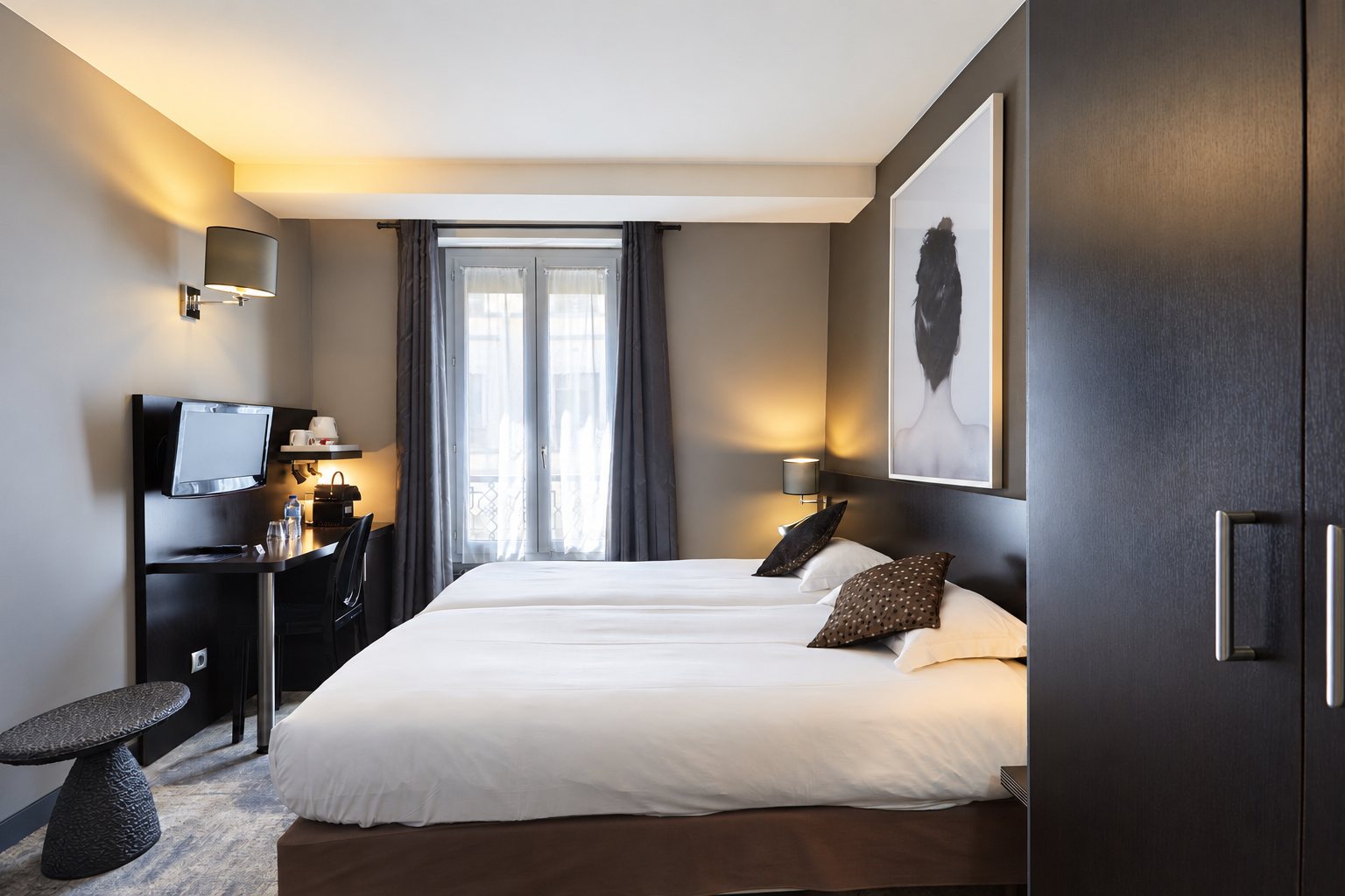 Chambre d'hôtel aux tons gris et bruns avec deux lits accolés, un bureau, une télévision et une grande photographie au mur.