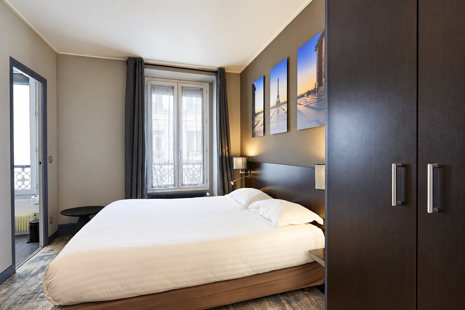 Chambre d'hôtel aux murs gris avec un grand lit blanc, une armoire sombre, une fenêtre drapée de rideaux et trois tableaux de Paris au-dessus de la tête de lit.