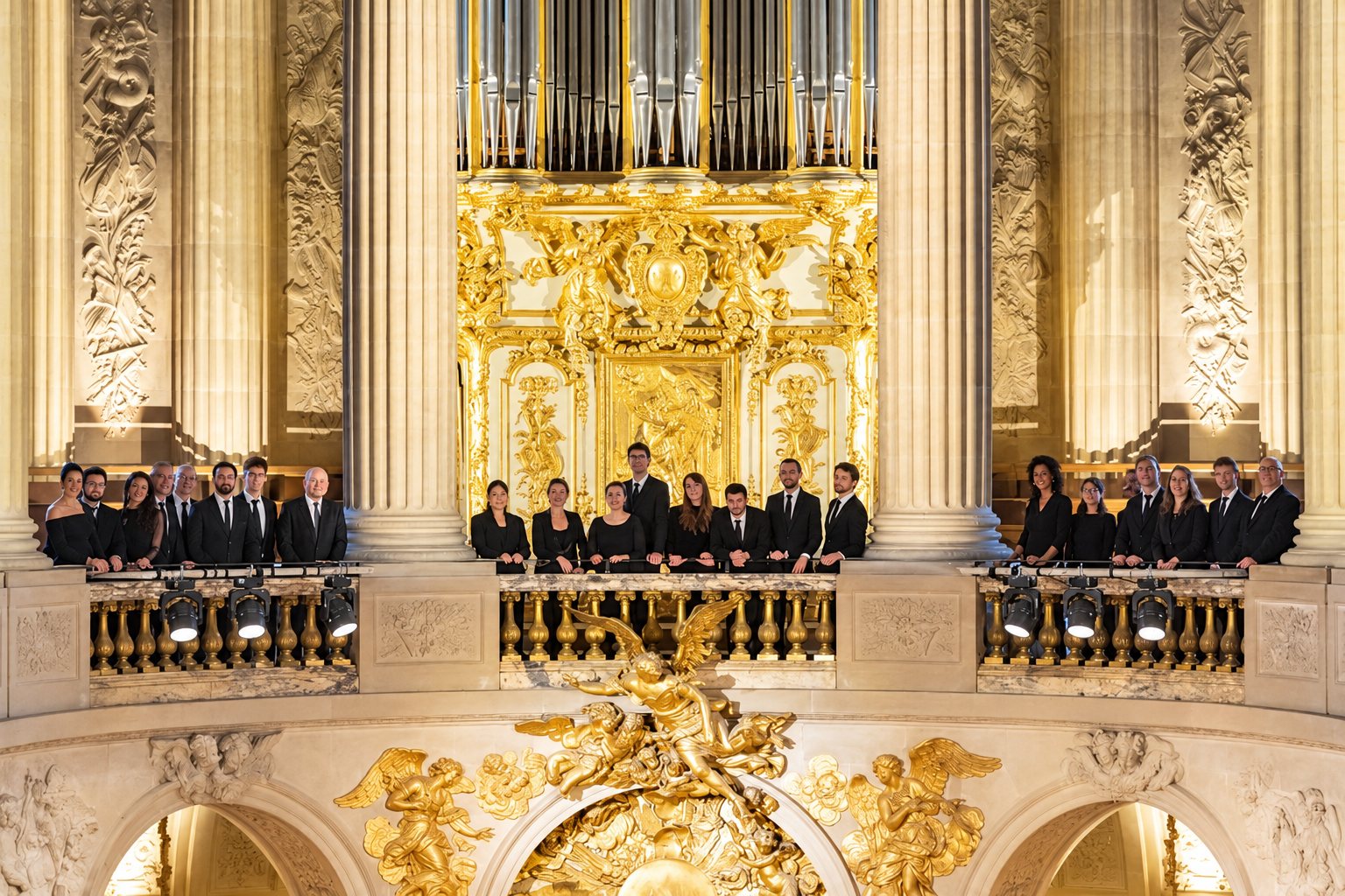 Un chœur vêtu de noir se tient sur une tribune devant un grand orgue aux décors dorés, entre de hautes colonnes de pierre et des sculptures d'anges dorés.