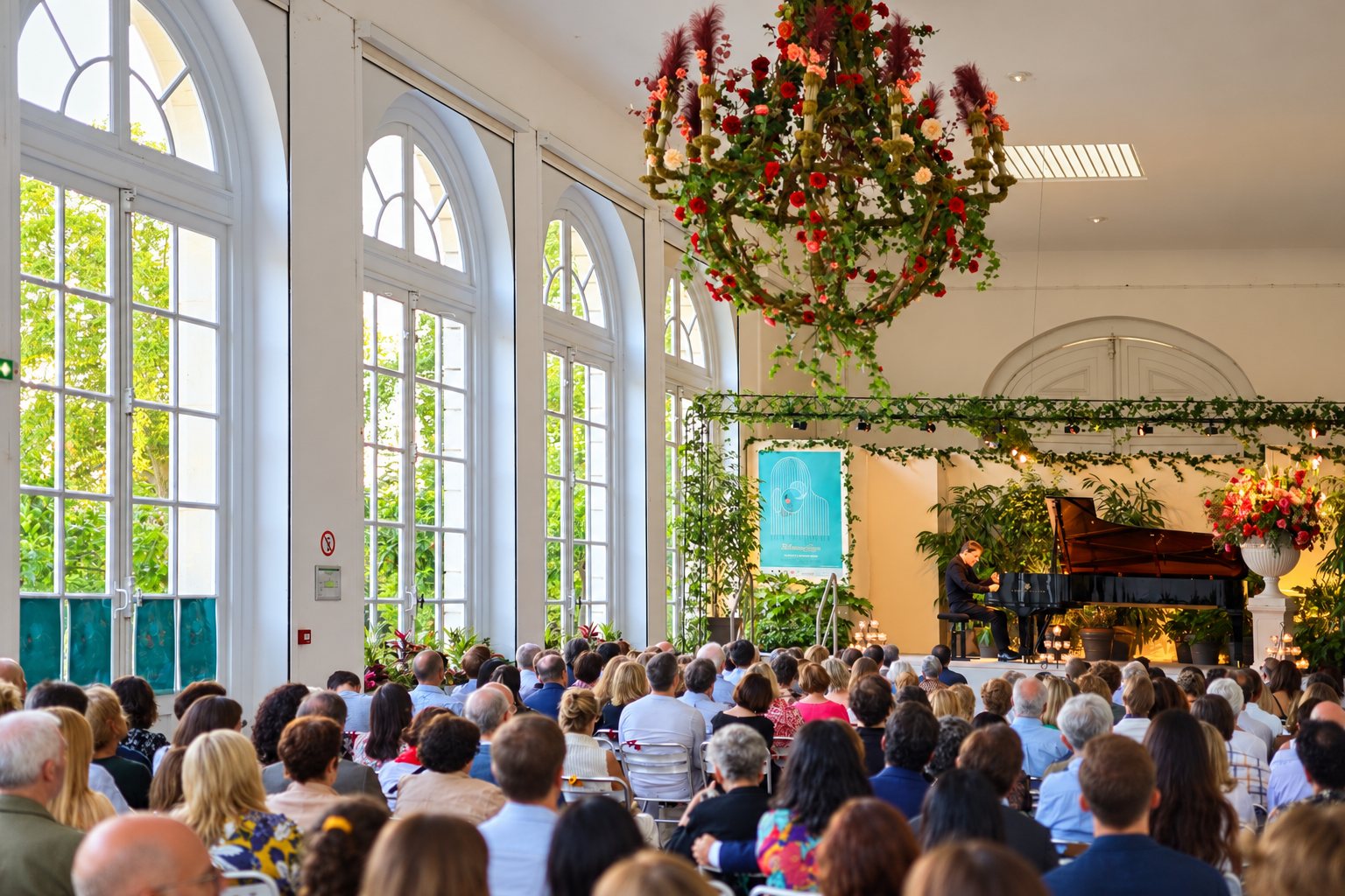 Un pianiste joue sur un piano à queue devant un nombreux public, dans une grande salle lumineuse aux hautes fenêtres cintrées et décorée de fleurs et de feuillage.