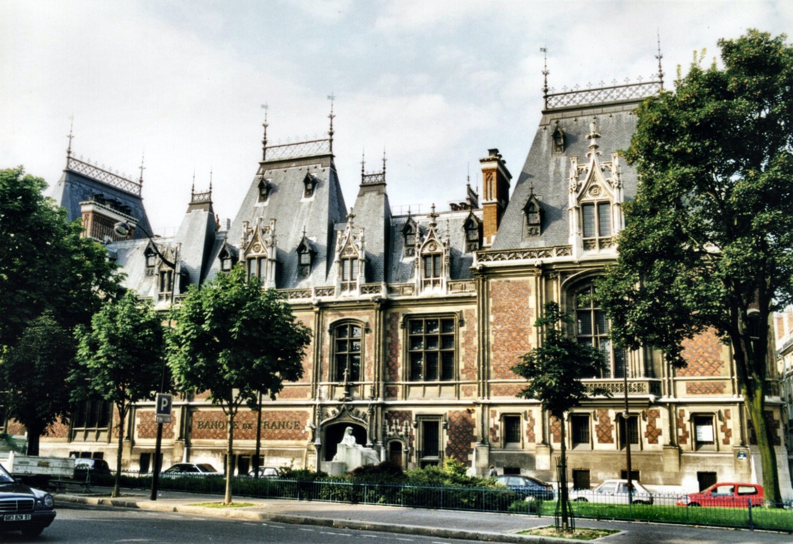 Façade d'un bâtiment néo-gothique en brique et pierre aux toits d'ardoise pointus, portant l'inscription Banque de France, avec des arbres et des voitures garées devant.