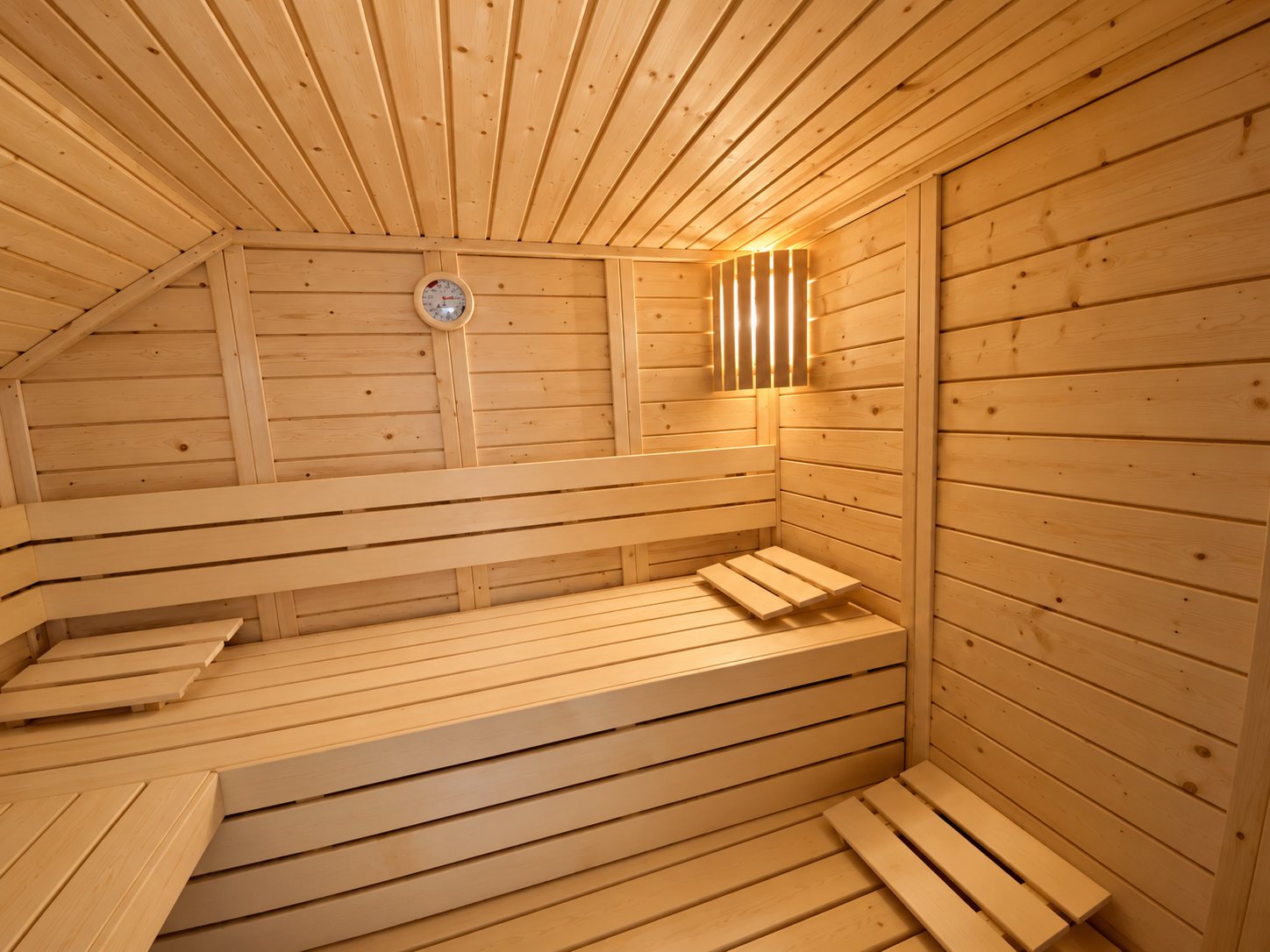 Intérieur d'un sauna en bois clair avec banquettes étagées, repose-pieds, hygromètre rond au mur et applique lumineuse à lattes.