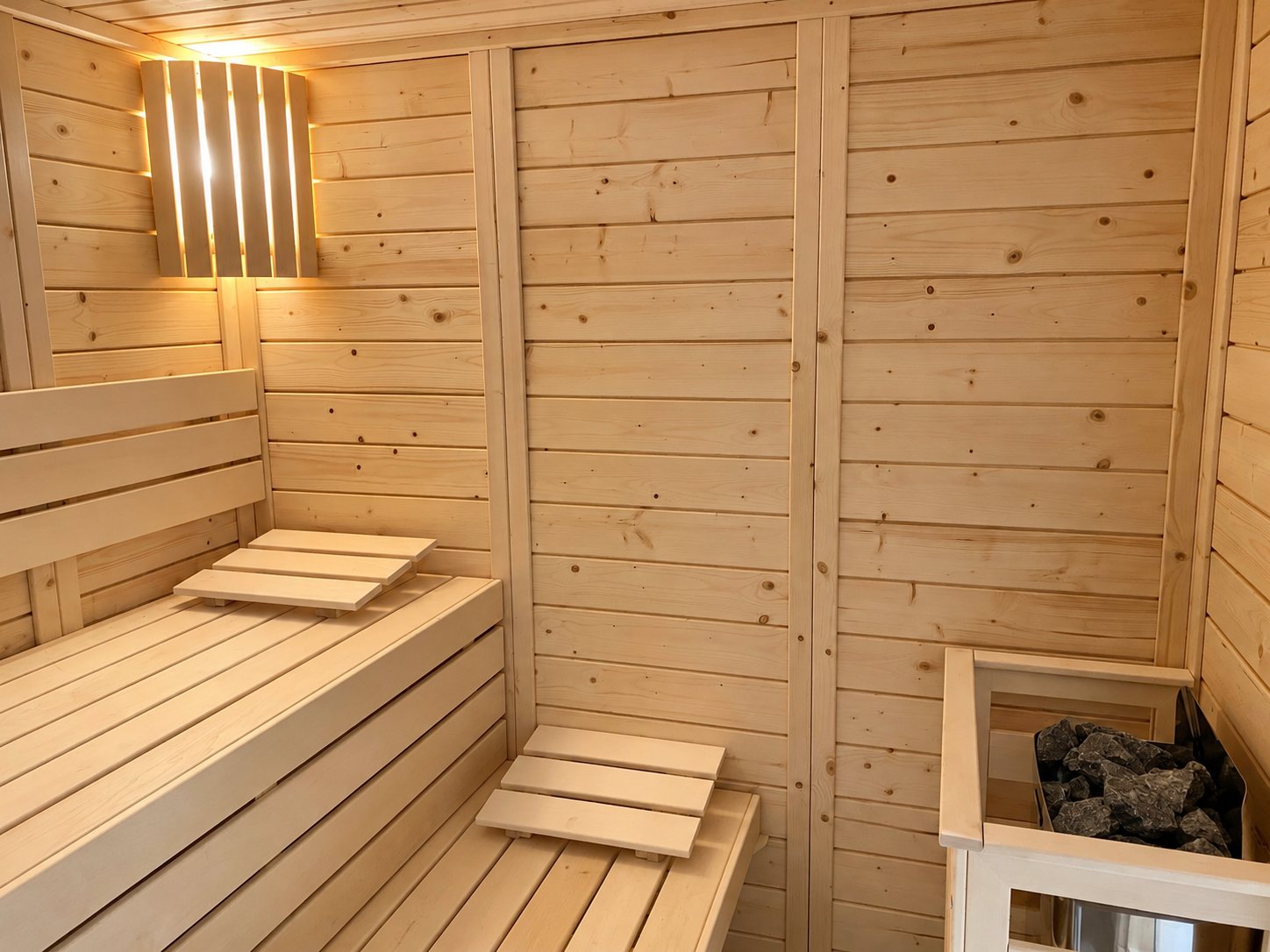 Intérieur d'un sauna en bois clair équipé de banquettes étagées, d'un poêle avec pierres et d'une applique lumineuse à lattes.