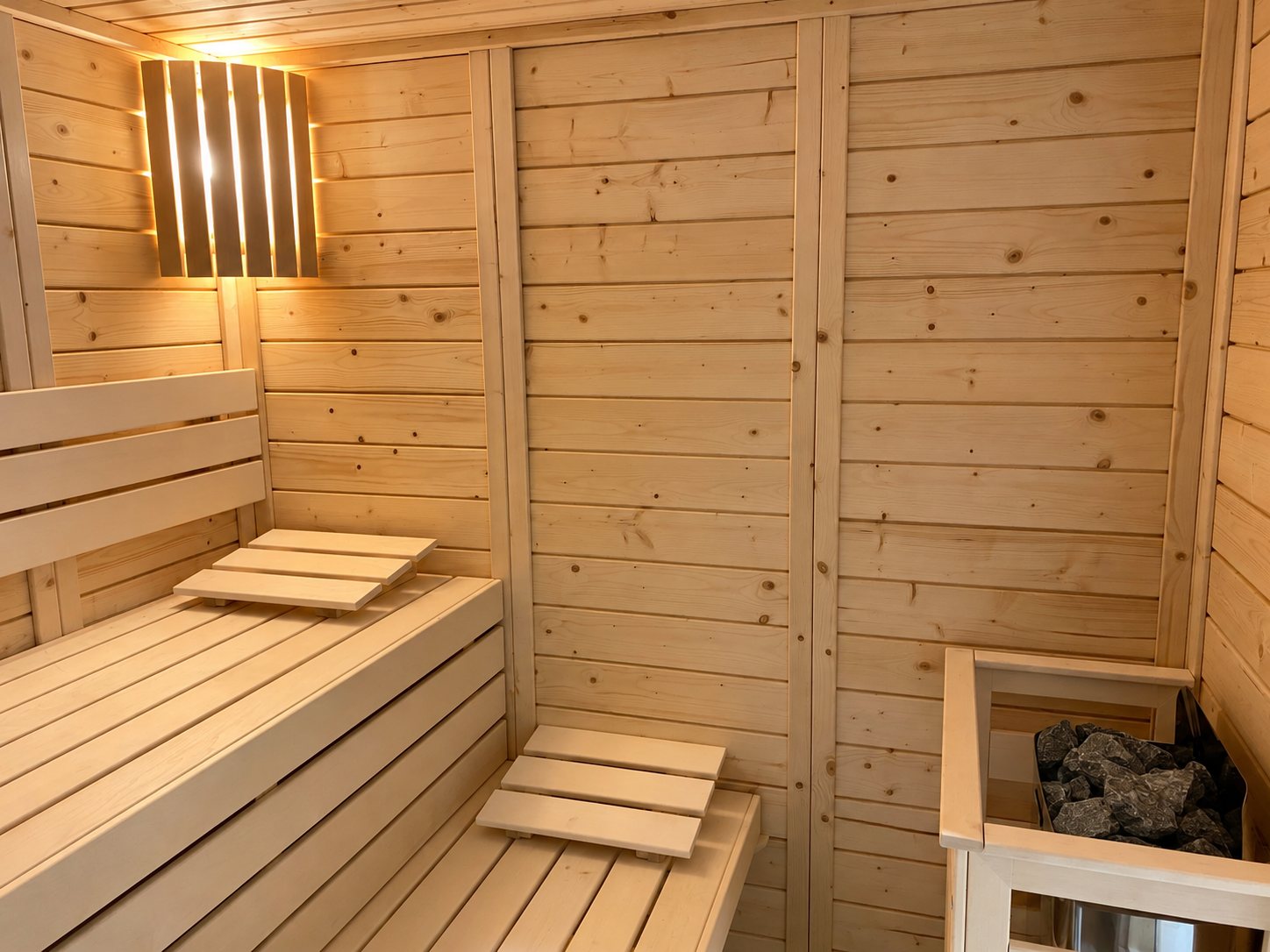 Intérieur d'un sauna en bois clair avec deux niveaux de banquettes, un luminaire à lamelles et un poêle garni de pierres.