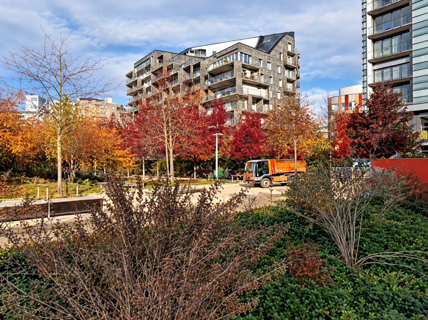 Un parc aux arbres aux feuilles rousses d'automne, avec un engin orange au loin et des immeubles résidentiels modernes à l'arrière-plan.