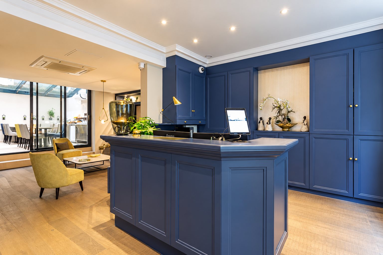 Hall d'accueil d'un hôtel avec comptoir et placards bleu marine, fauteuils jaunes, parquet clair et salle de petit-déjeuner vitrée au fond.