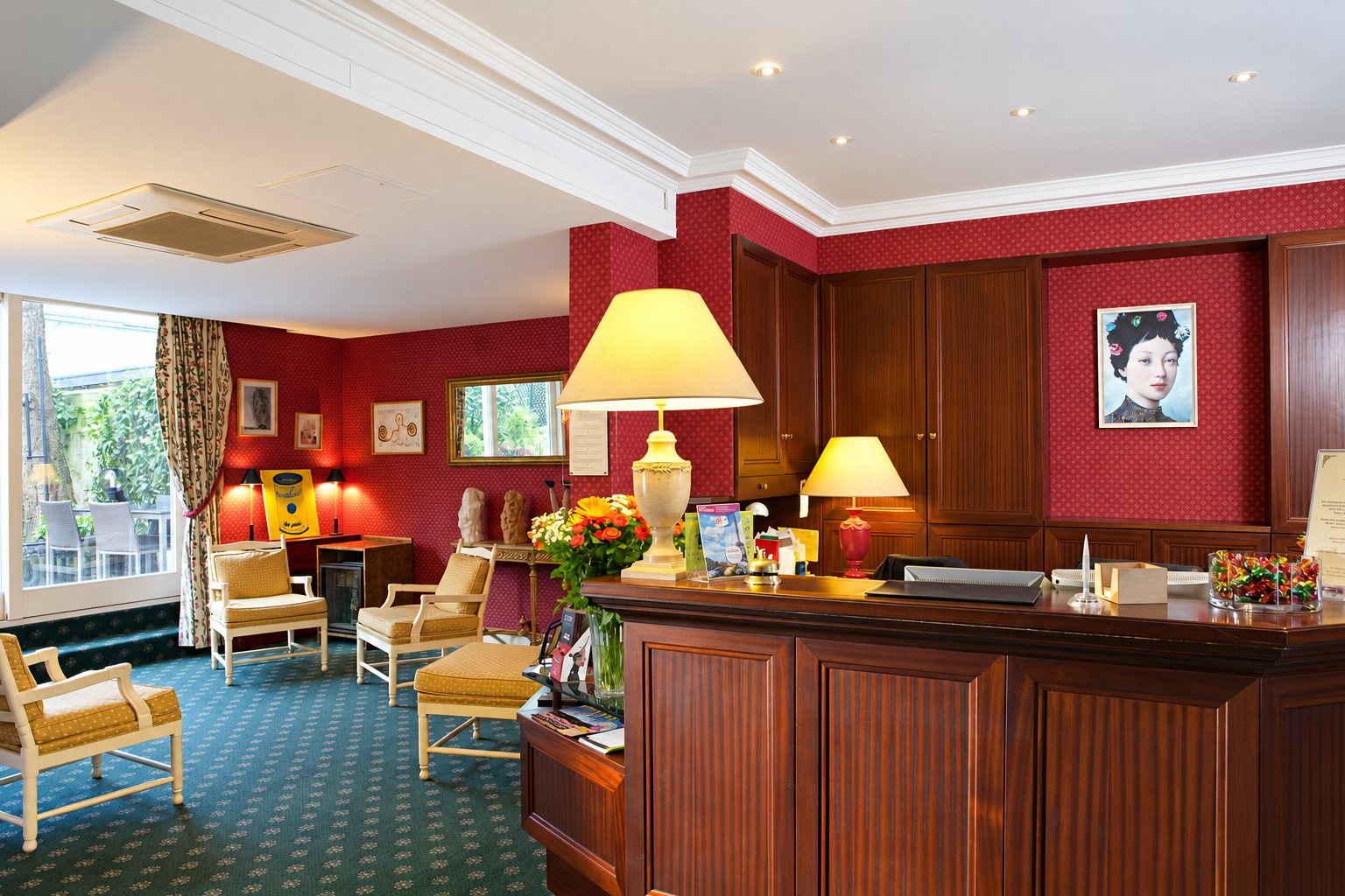 Réception d'un hôtel avec comptoir en acajou, lampes allumées, bouquet de fleurs et fauteuils dans un salon aux murs rouges.