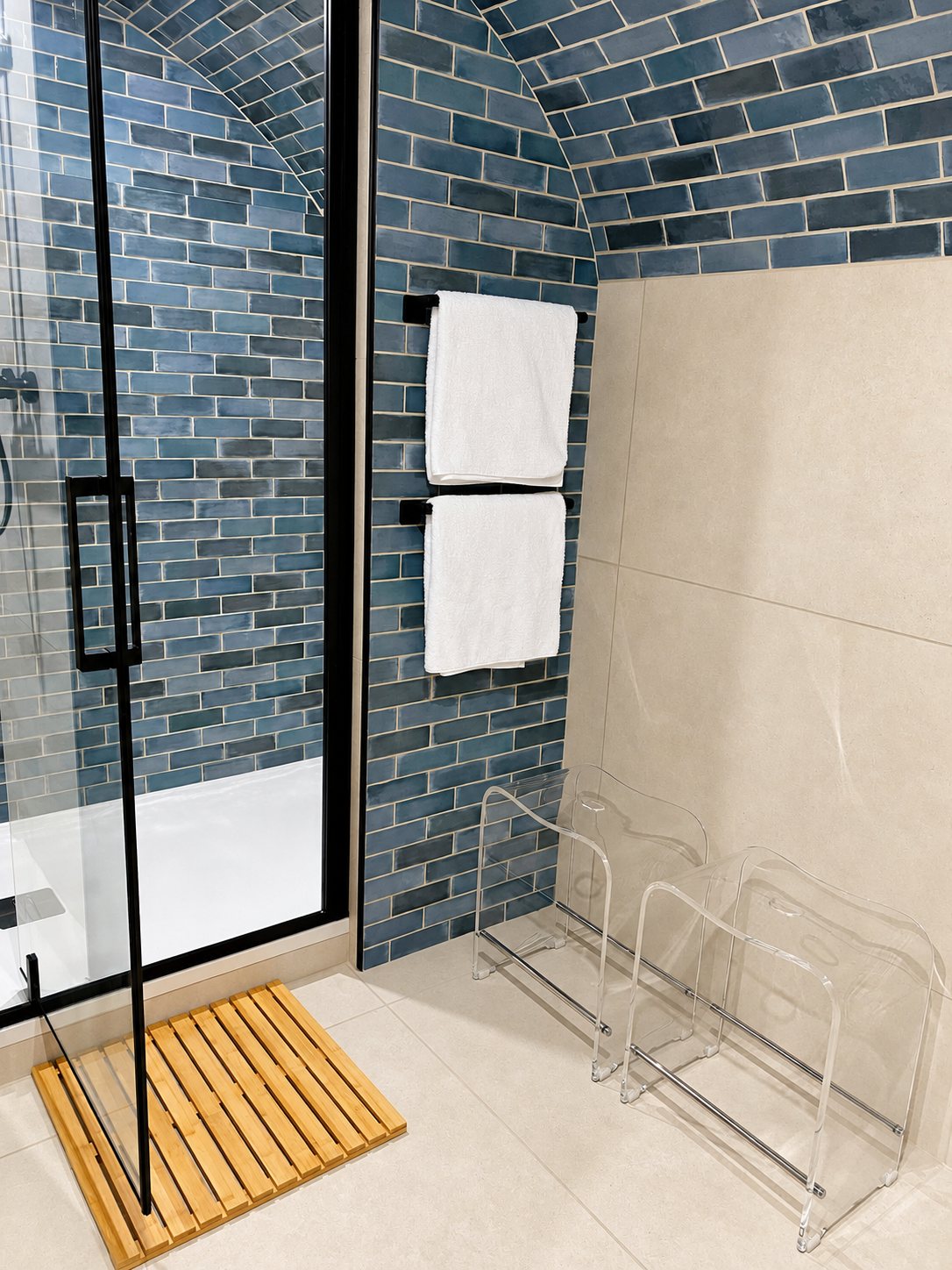 Salle de bain aux murs voûtés en carrelage bleu, avec douche vitrée à cadre noir, serviettes blanches, tabourets transparents et tapis en bambou.