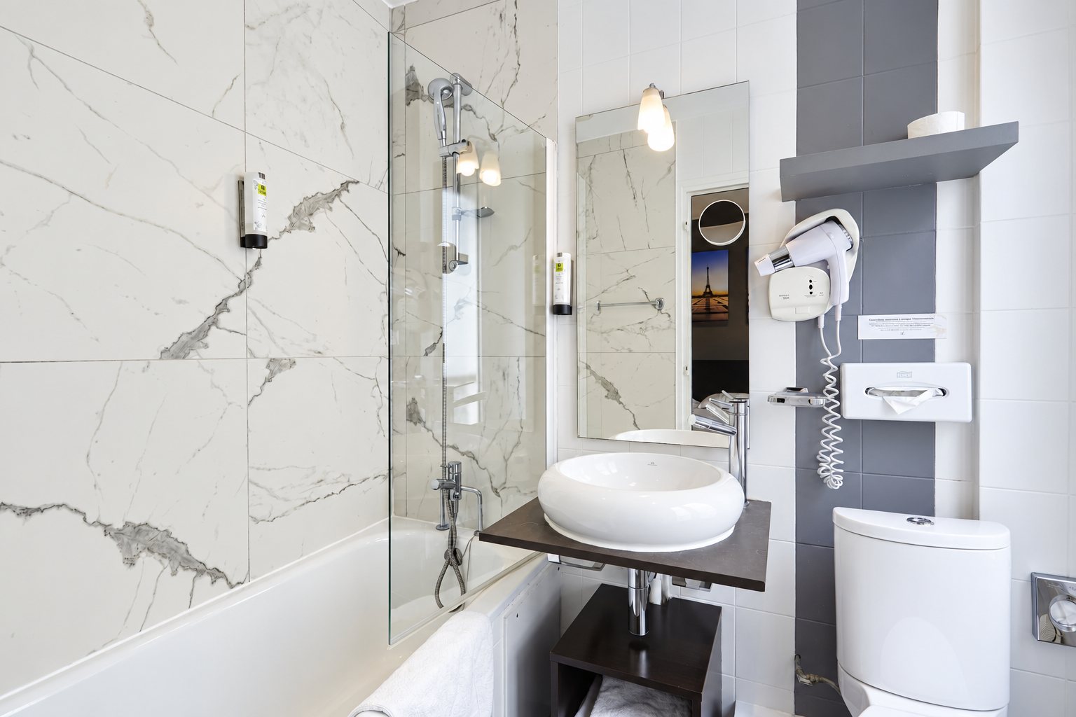 Salle de bain lumineuse d'hôtel avec murs en marbre blanc, baignoire avec pare-douche en verre, vasque ronde posée sur une tablette, sèche-cheveux mural et toilettes.