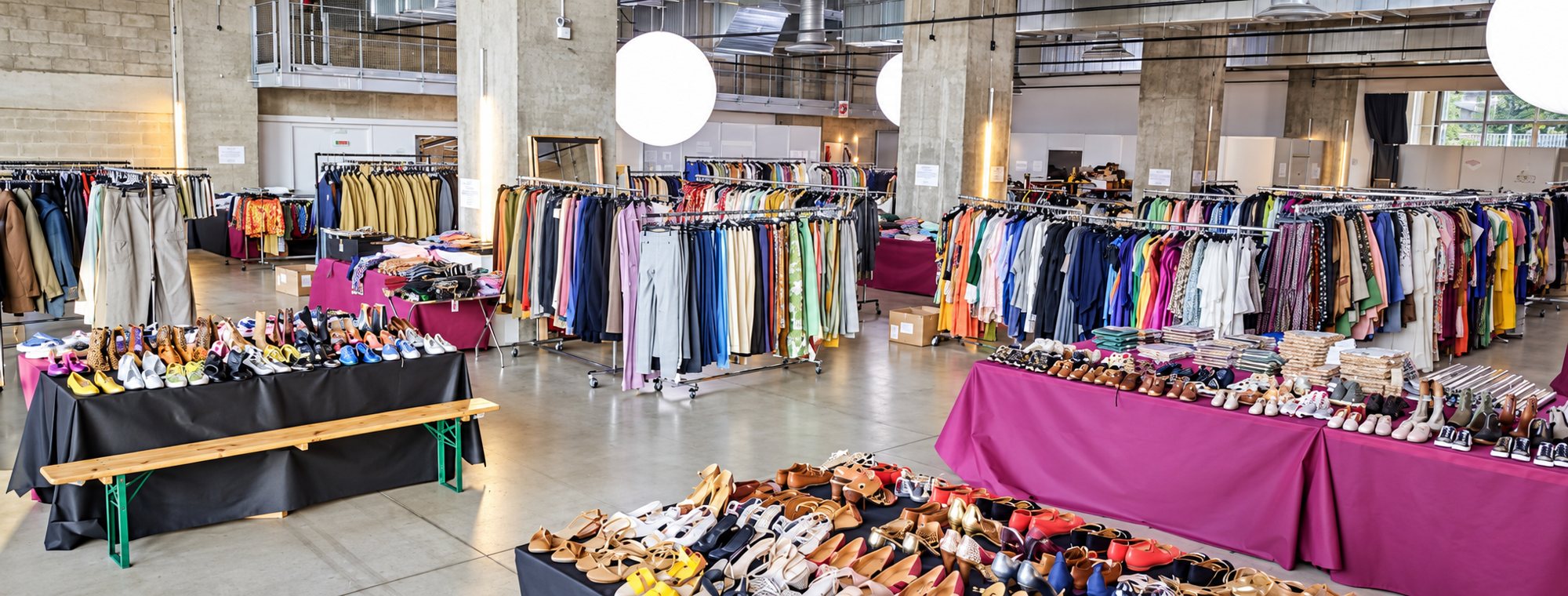 Vaste espace au sol en béton où sont présentés des portants de vêtements colorés et des tables couvertes de chaussures de toutes sortes.