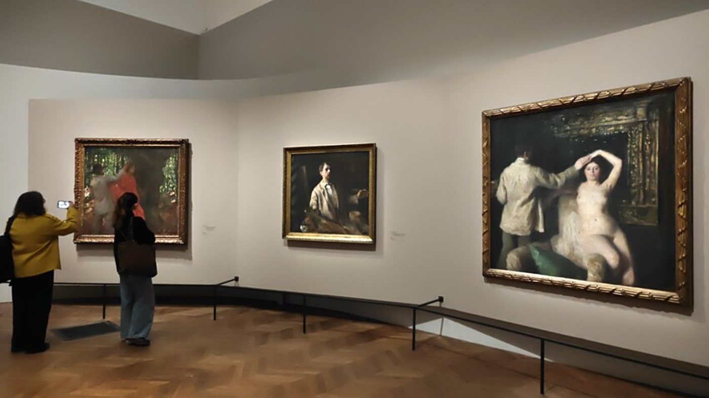 Dans une salle de musée au parquet en chevrons, deux visiteuses regardent et photographient trois grands tableaux dans des cadres dorés accrochés aux murs clairs.
