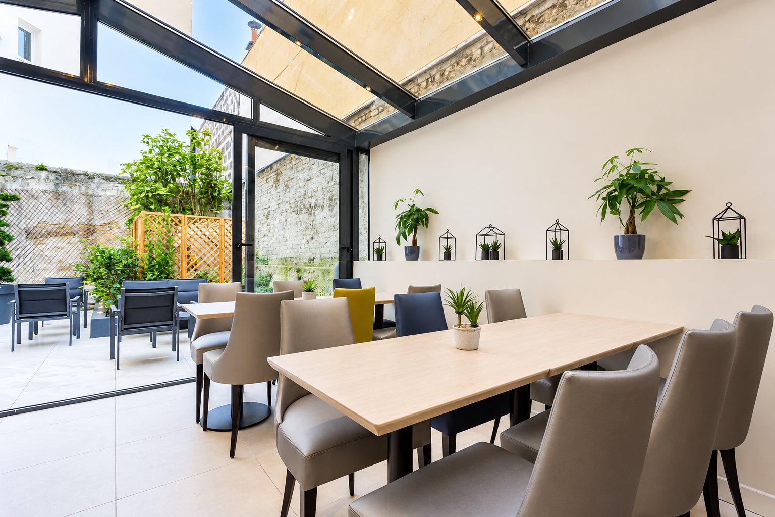 Salle à manger claire sous une verrière, avec tables en bois, chaises beiges, plantes sur une étagère et une terrasse en pierre à l'extérieur.