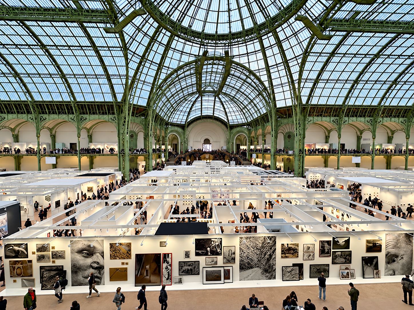 Vue en hauteur d'un salon d'art installé sous une immense verrière, avec des stands blancs présentant des photographies et de nombreux visiteurs.