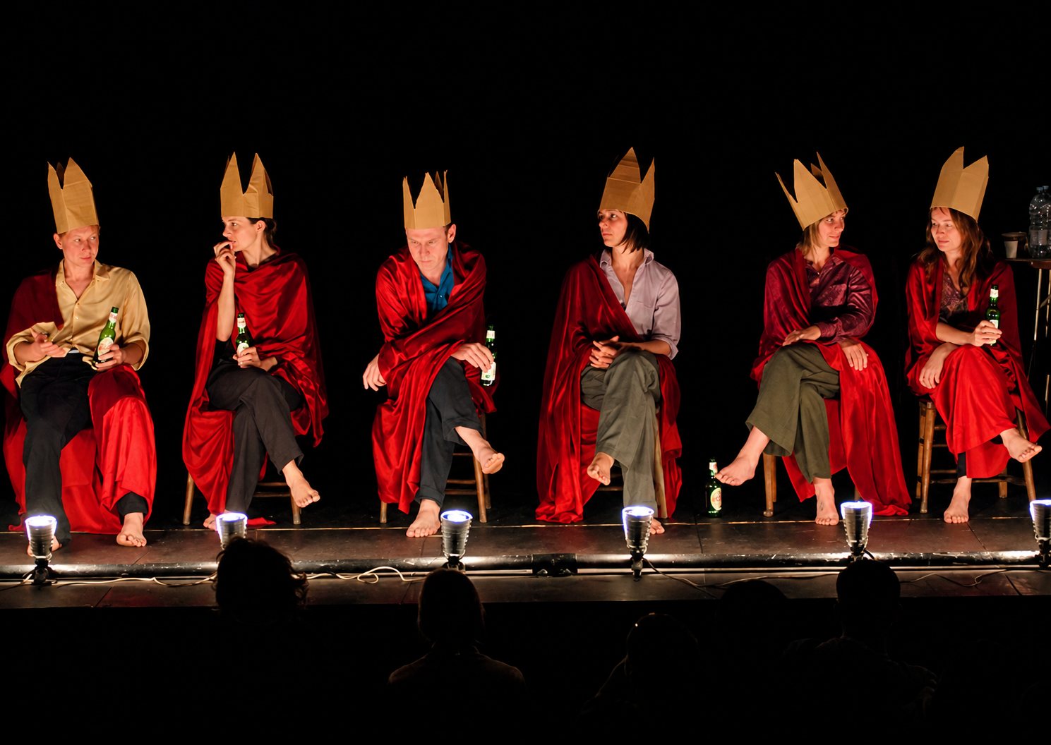 Six comédiens pieds nus, assis côte à côte sur une scène sombre, portent des capes rouges et des couronnes en papier doré, certains tenant une bouteille de bière, devant le public en silhouette.