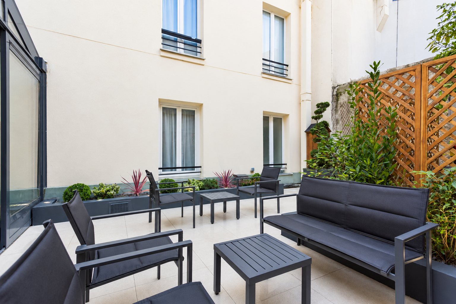 Une cour d'immeuble aménagée en terrasse avec un canapé, des fauteuils et des tables basses gris anthracite, entourés de plantes et d'un treillis en bois.
