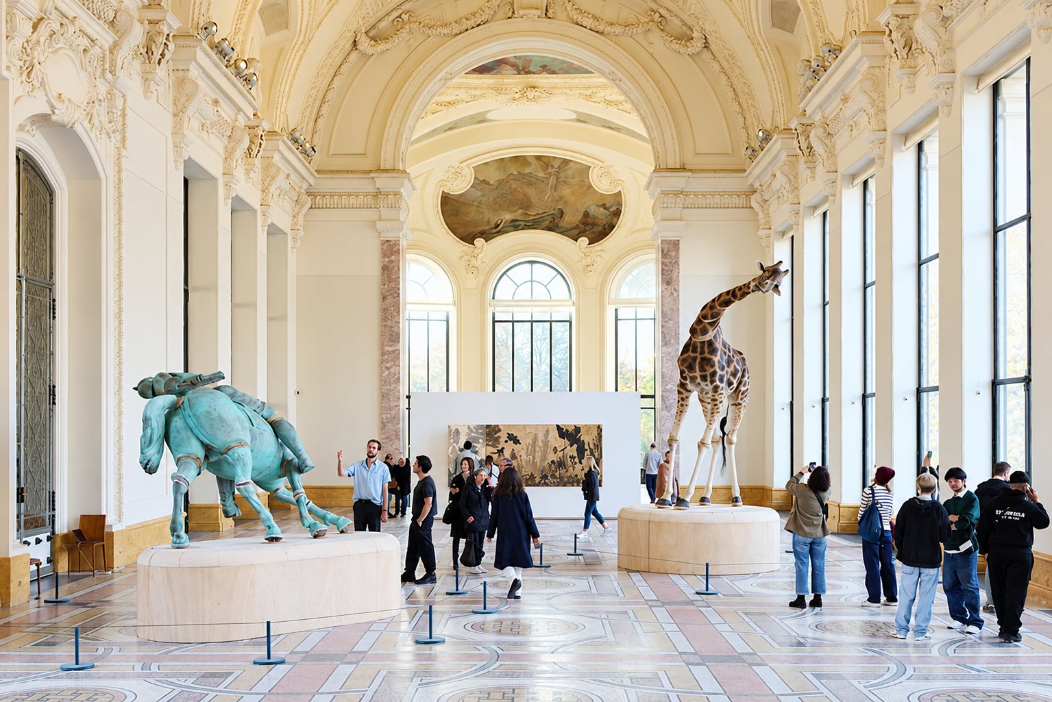 Des visiteurs parcourent une grande salle de musée aux décors dorés où sont exposés une girafe naturalisée et une sculpture de cheval en bronze vert-de-gris.